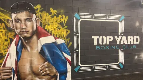 Richard Knights/BBC A mural is on a wall of the gym which has a picture of Amir Khan on the left with a United Kingdom flag around him. On the right of the mural it says 'Top Yard Boxing Club' in white and blue writing.