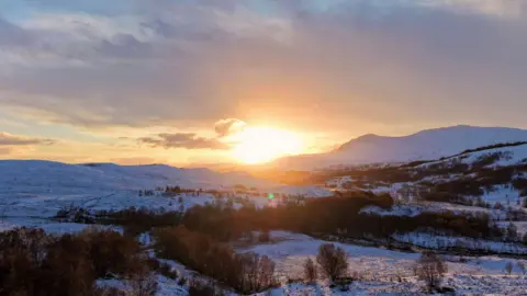 Andy Bugeja A peaceful winter landscape with rolling snow-covered hills, scattered trees, and a warm golden sunset casting soft light across the scene.
