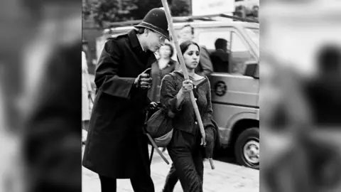 During a protest, a young woman holds the stick of a placard while a police officer in a black coat and helmet looks like he is communicating something to her while leaning in and gesturing with his hand. She looks very focussed on moving forward. She is wearing dark clothing with a cardigan.