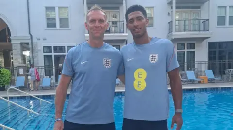 Chris Walton Walton is on the left, next to Bellingham on the right. They are both wearing light blue tops including the coat of arms for the England national team, which features three blue lions. A swimming pool and a building are behind the men.