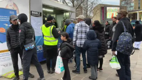 Aisha Iqbal/BBC A queue of people outside a food truck. It is in a city square, with a big screen and shop frontage visible in the background.