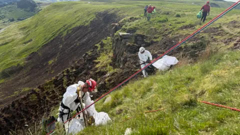 Central Beacons Mountain Rescue Team Close up of volunteers in white uniform attached to red ropes on the mountain clearing the rubbish. 