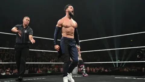 Getty Images A male wrestler is standing in the ring. He is wearing navy trousers and navy arm covers over half his arm. The referee is behind him. The outline of another wrestler is also behind him. 