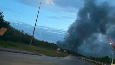 Wide view of a large black cloud of smoke rising in the air behind a road. The sky is dark as the sun begins to rise.