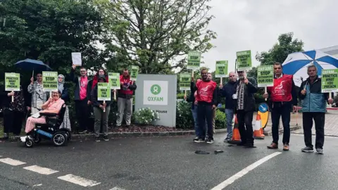 Unite A group of about 15 protesters gathered in the car park. Placards say things like 'Hey Oxfam walk the walk' and 'Oxfam: stop outsourcing our jobs'.