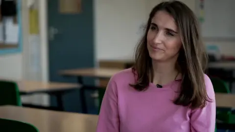 A woman with long brown hair and a pink top sits in an empty classroom