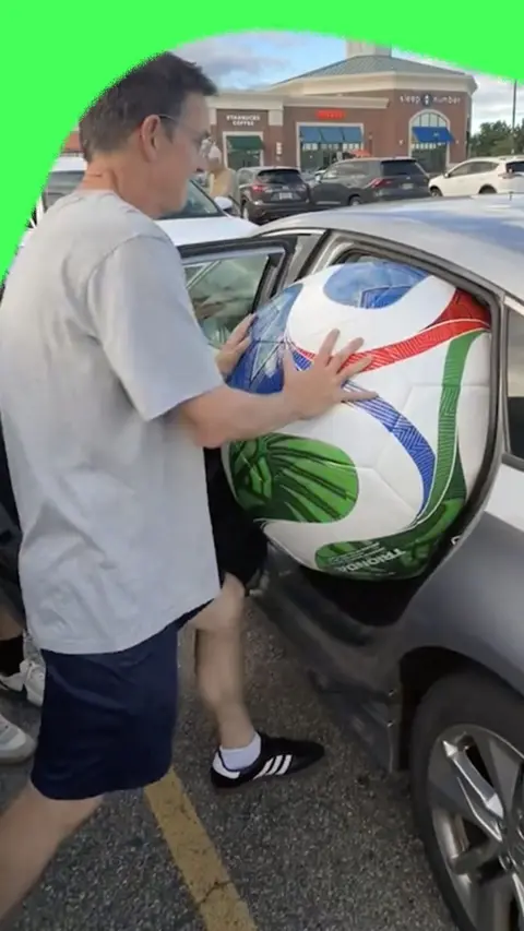 Kevin Cronin, wearing a grey t-shirt and navy blue shorts, puts a giant Fifa World Cup Football in the back of his car