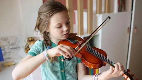 Getty Images A stock image of a little girl using a violin inside a room. She is wearing a teal green top and has colourful bracelets on. The background is out of focus but a white cabinet can be seen behind her.