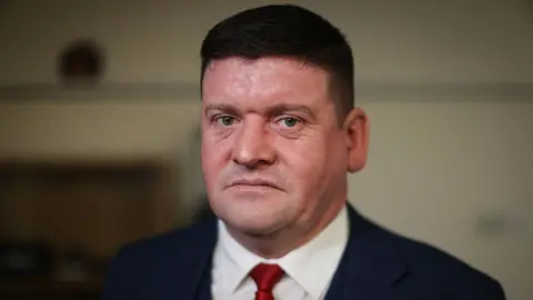 A man in his 40s with short dark hair and wearing a blue suit and red tie