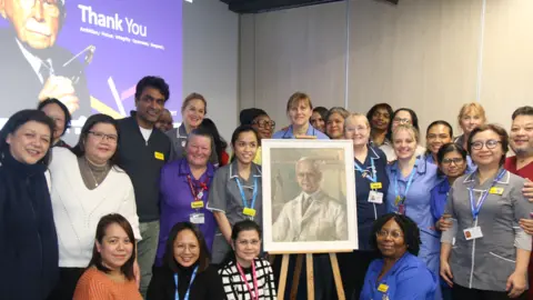 Guy’s and St Thomas’ NHS Foundation Trust Staff from Guy’s and St Thomas’ NHS Foundation Trust gather round an easel displaying a portrait of Sir Harold Ridley. In the background a projected image on the wall features another image of him with the words 'thank you'