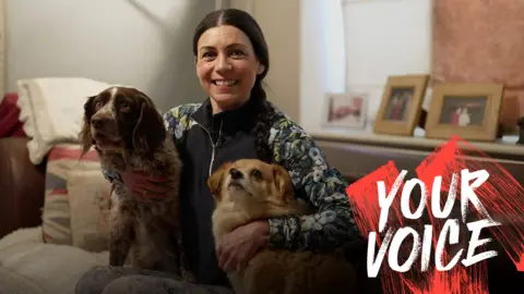 Helene Svinos sits on a sofa in between two dogs, with her arms around them. She is smiling and wearing a dark gilet over a patterned top, and leggings, with her dark hair tied back. There are cushions on the sofa and photo frames in the background.