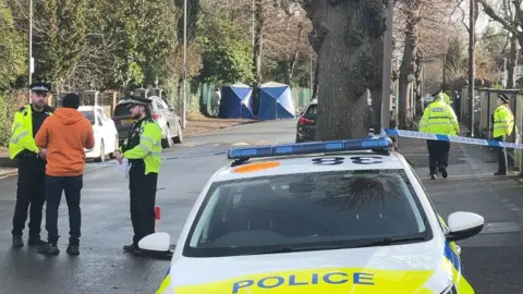 Addington Road, a tree-lined street in Reading. In the foreground is a police car. Two police officers stand near it talking to a member of the public. In the background are two blue police forensics tents.