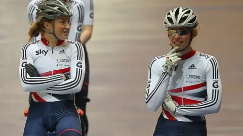 Getty Images Vicky Williamson is wearing a white, red and blue all-in-one lycra outfit and cycling helmet and chatting to her teammate beside her. She is on her bike but has her arms crossed.
