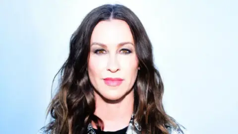 Shelby Duncan/Lytham Festival Alanis Morissette stands against a light blue background. She has long wavy dark hair and is wearing a black and white collared shirt.