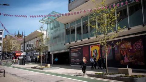 A street with the glass front of the Mander centre on the right with a blue sky behind and Union Jack bunting. The back of a shopper can be seen and a group of several people are walking past a sign with colourful artwork advertising Wolverhampton Art Gallery. 