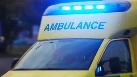 A ambulance pictured with its blue lights on. The word "ambulance" is written on the hood over the windscreen.