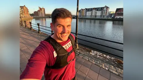 Scott Lawther A man with short brown hair wearing a red tshirt and a black running vest, he is smiling at camera as he takes a selfie, running alongside the water in Belfast. There are high rise waterfront apartments visible on the horizon.