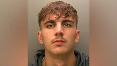 A mugshot of Kian Rowe. He has short brown hair, shaved on the sides and longer on the top, with some strands of hair coming down on to his forehead. He has blue eyes and a sparse moustache. He is wearing a dark grey hoodie and is looking serious.