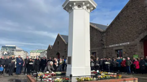 Gemma Daubeney/BBC People gather outside the Maritime Museum where a number of wreaths have been laid beside the tall white memorial. An older woman is placing her wreath there, whilst others who are holding floral tributes look on.