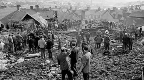 A black and white image of the aftermath of the Aberfan disaster. There is rubble everywhere, damaged houses and men looking at the carnage caused.