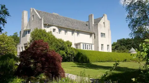 National Trust for Scotland Charles Rennie Mackintosh's house before the box was put around it. The white walls sits in the midst of a green garden. The sun shines around the house. 