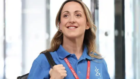 Natalie Cassidy stands in a building, smiling, wearing the blue polo shirt of North Hertfordshire College and carrying a black rucksack