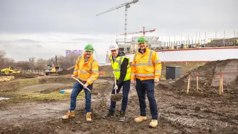 Sunderland City Council (L-R) Andy Radcliffe, chief executive of Esh Construction, councillor Kevin Johnston and Stephen McClean, construction manager at Esh Construction at the building site of the new park. Mr Radcliffe and Johnston are both holding spades which have been used to dig up earth. Excavators can be seen in the background and the ground is muddy.