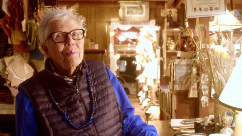 Ryan Louie Sue Johnson, owner of Sue Johnson Lamps in Berkeley, California has short white hair and glasses. She is wearing a puffy vest, blue shirt and necklace and is surrounded by different kinds of lamps