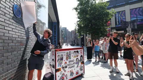 Brad Kella is standing, and pulls a white cover from a blue plaque as people watch and take photographs on their phones beside a decorated piano. It is sunny day. 