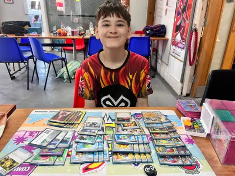A young boy with black hair smiles at the camera surrounded by his pokemon cards