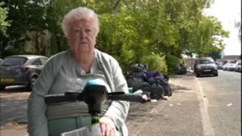 A woman with short white hair sits on a mobility scooter on a pavement. Behind her is a pile of black bin bags.