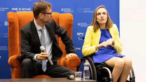 Lucy Robinson during an event. She is a wheelchair user. She has a head mounted microphone and is looking to her right, away from the camera. She is wearing a blue dress and a yellow jacket. A man holding a microphone is sitting in a brown armchair next to her. He is looking at her. There is a bottle of water and a glass on the table in front of them.