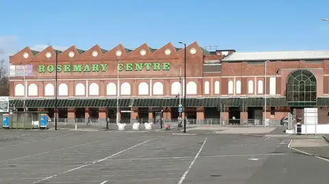 LDRS A large brick commercial building with filled in windows, glass portico and extensive car park in front