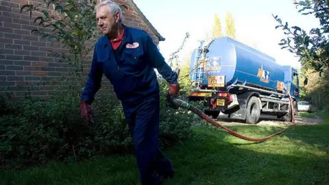Getty Images Tanker driver Bill Webb, dressed in blue overalls and carrying a large pipe that is connected to the tanker, delivers domestic heating oil to a house in Newbury in 2007.