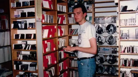 Ronan Munro A young skinny Ronan working in Our Price. He wears a white band t-shirt tucked into jeans. Picture vinyl displays of Bon Jovi and The Cure are behind him.