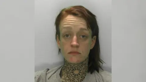 Gloucestershire Police Mugshot of Hannah Roberts - a woman in her early 30s with a distinctive, tribal-style tattoo covering her neck. She has medium length dyed red hair, and is wearing a grey tracksuit.