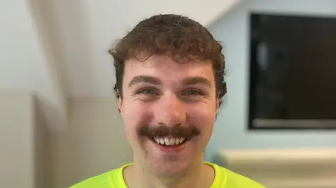 Steve Jones/BBC Bailey Greetham, que tiene el pelo y el bigote castaños. Aparece sonriendo en la foto de primer plano.