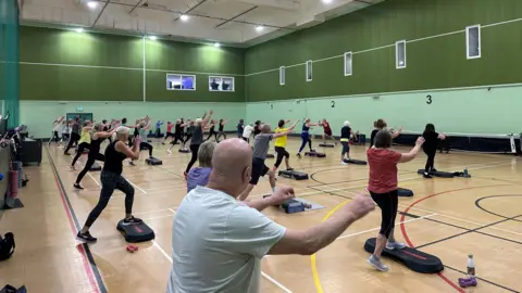 A sports hall filled with people in sports wear with their hands in the air as they use a black step as part of their workout.
