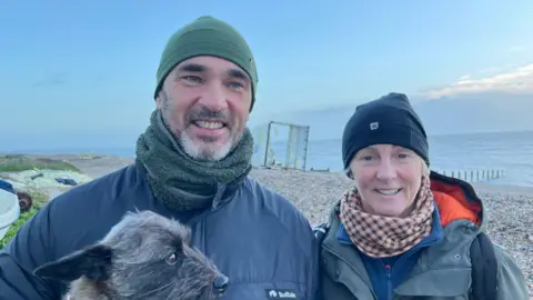 A man and a woman in winter clothing stood side-by-side on a pebble beach looking and smiling at the camera. behind them is the sea and a wrecked shipping container washed onto the shore