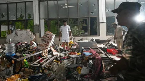 People stand amid a chaotic scene of debris within the doctors' mess hall after the crash. It is clearly dark outside, meaning it is several hours since the incident