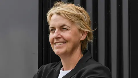 SNS Leanne Dempster has short blonde hair and smiles while looking off to one side. She wears a white t-shirt and a black blazer