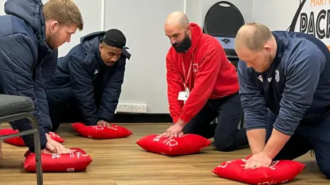 Empat pria berada di sebuah ruangan, berlutut, masing -masing mensimulasikan kompresi dada pada bantal merah. Tiga dari mereka berada di Cardiff Rugby Training Kit. Pria keempat mengenakan jumper Yayasan Jantung Inggris Merah. Lantainya kayu dan di latar belakang ada dinding putih dengan kursi bertumpuk dan soket steker.