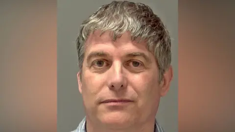 A police custody image of a man who is looking directly at the camera and slightly smiling. He has grey hair that slightly hangs over his forehead and he is wearing a blue shirt with white stripes on it. 