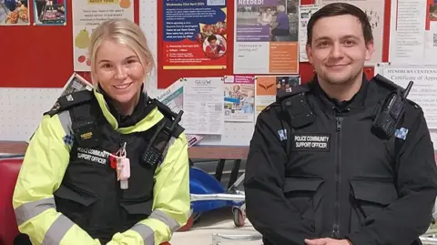 A female and male police officer, in uniform, sitting at a table with posters in the background during community engagement event