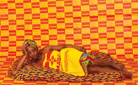 Adji Dieye A woman lies on a soft blanket, looking into the camera, wearing Maggi stock cube branded dress, behind her a wallpaper made of the Maggi logo