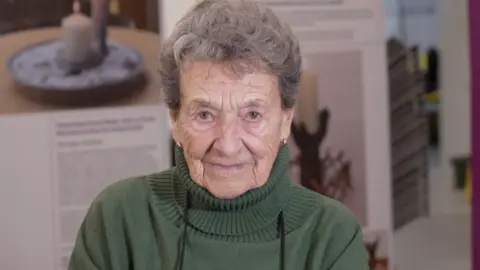 Hedi Argent sits facing the camera, wearing a green jumper, with display boards behind her.