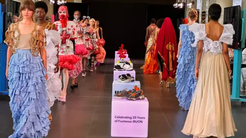 Carroll Weston/BBC Models walk up and down a catwalk at the University of Northampton. They are wearing bright colours and in the centre of a room are plinths displaying different shoe and trainer concepts.