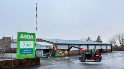 The front of a supermarket, with extensive damage to an entrance foyer. There is a green totem at the front of the car park and a red telehandler located a few metres away from the entrance. The rest of the building appears undamaged.
