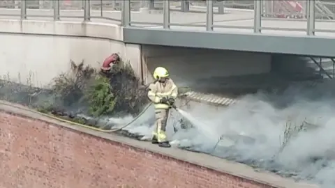 A firefighter, in protective clothing, stands on a wall and sprays water onto a smouldering fire with grey smoke. The grey structure of a bridge is visible in the background.