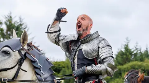 HES A man dressed as a medieval knight punches the air and shouts to the crowd. He is on horseback on a white horse